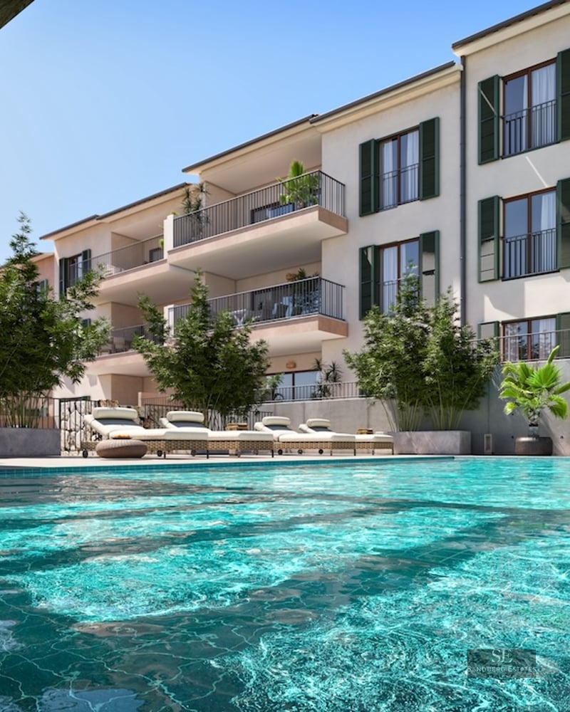 Large turquoise swimming pool with sun loungers in front of an apartment building with green shutters and balconies.