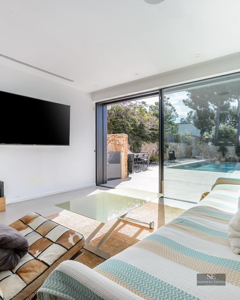 Bright living room with a striped sofa, large TV, and floor-to-ceiling glass doors leading to a pool terrace.