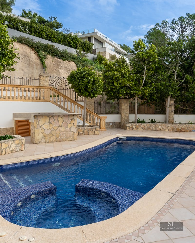 Blue swimming pool with integrated seating on a spacious tiled terrace surrounded by balustrades and trees.