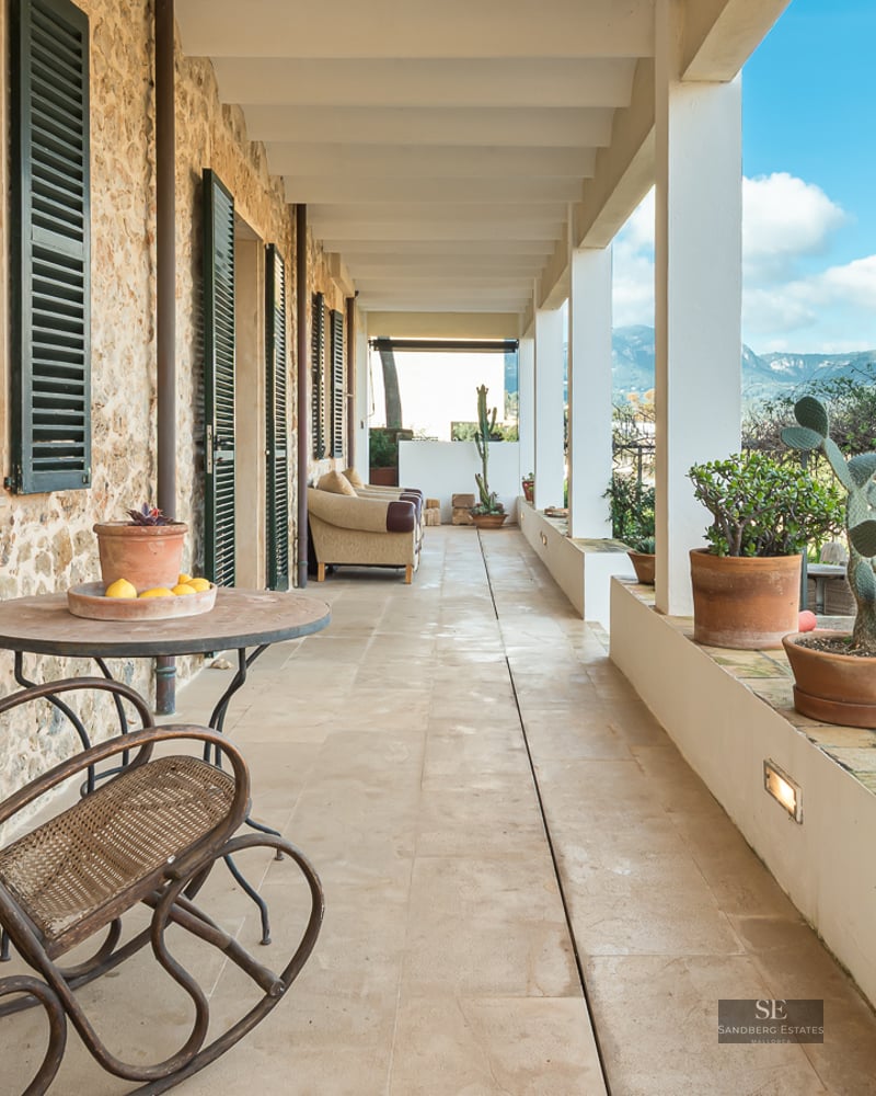 A long covered stone terrace with terracotta pots, green shutters, and a rocking chair overlooking mountains.