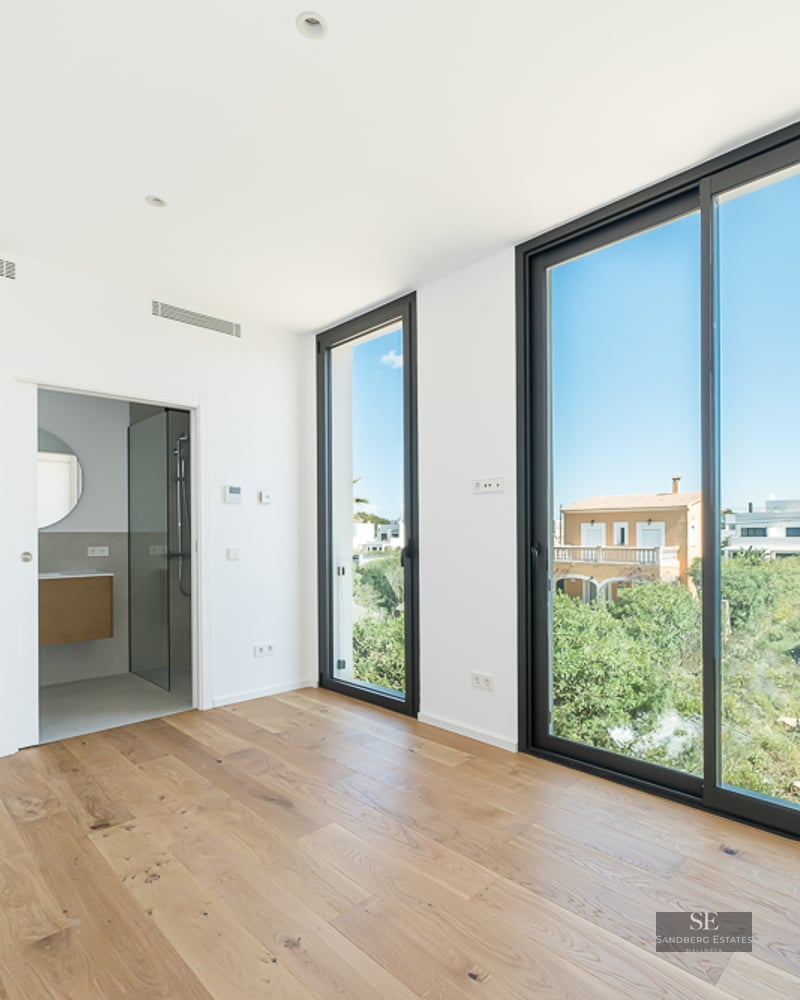 Empty modern bedroom with light wood floors, large black-framed windows overlooking greenery, and an open ensuite bathroom.