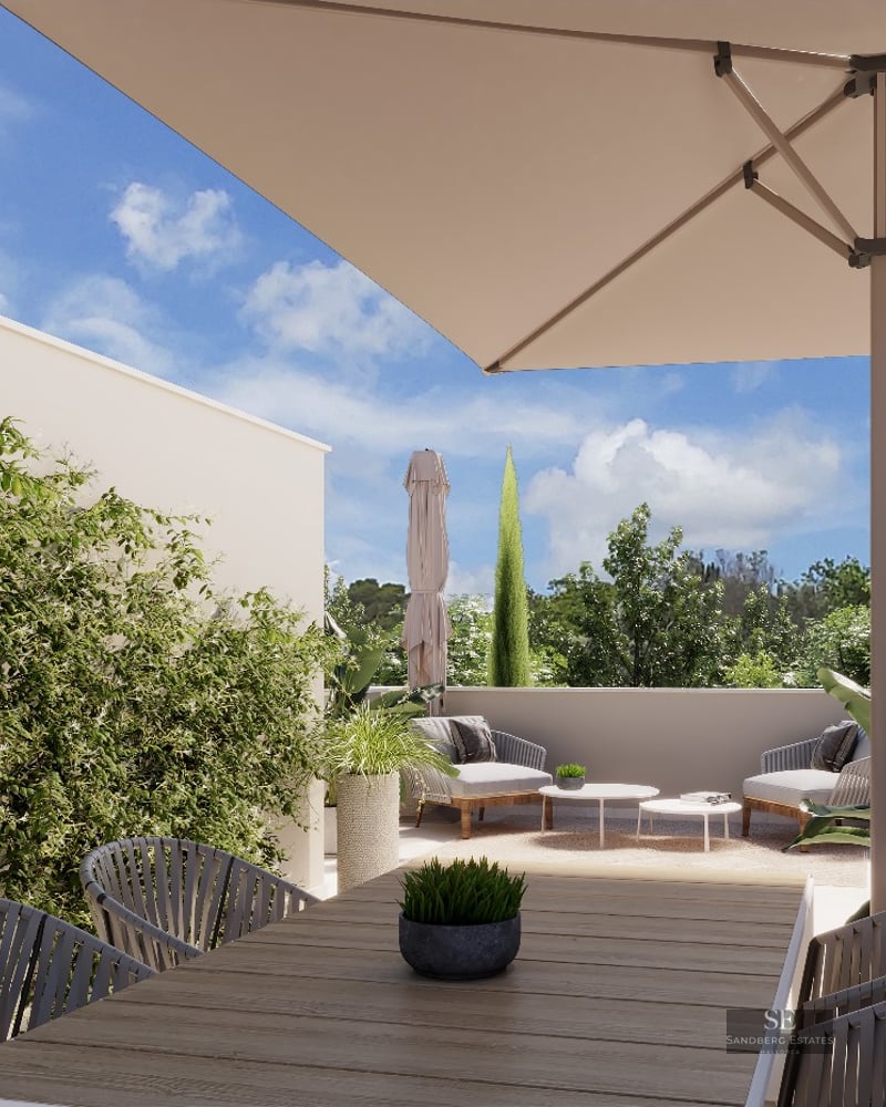 Bright outdoor terrace with a wooden dining table, large sun umbrella, lounge chairs, and lush green plants.