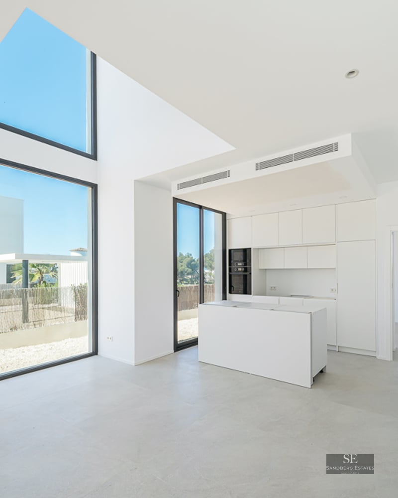 Minimalist white kitchen with an island, double-height floor-to-ceiling windows, and bright natural light.