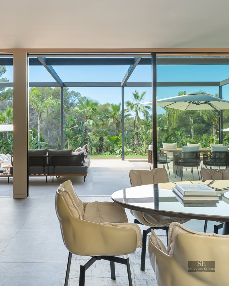 Modern dining room with a marble table and linear fireplace, opening through glass doors to a lush tropical terrace.