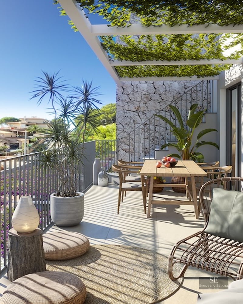 Sunny modern terrace with a wooden dining table, wicker lounge chair, and a plant-covered pergola overlooking a coastal town.