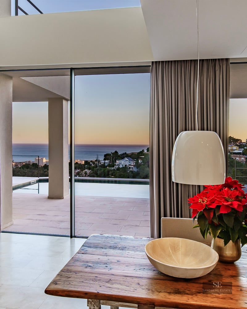 Interior dining area with a rustic wooden table overlooking an outdoor terrace with an infinity pool and sea view.