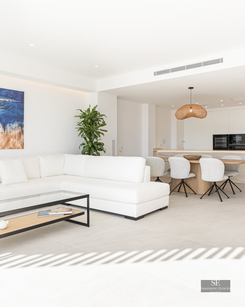 Bright living room with white L-shaped sofa, glass coffee table, abstract art, and integrated kitchen and dining space.