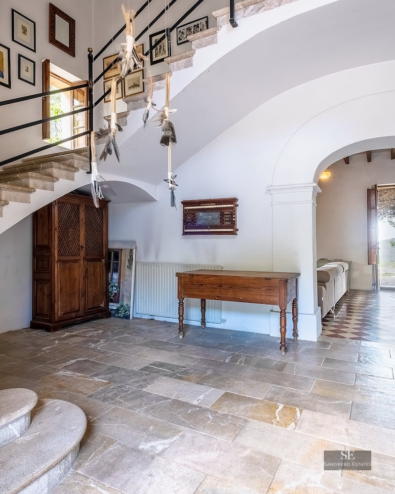 A grand entrance hall with a stone staircase, antique wooden furniture, and a gallery wall under an arched doorway.