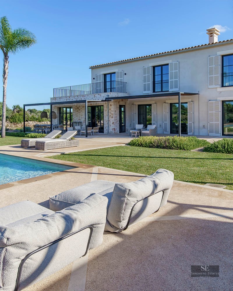 Large turquoise swimming pool in front of a stone villa with palm trees and green lawn under a clear blue sky.