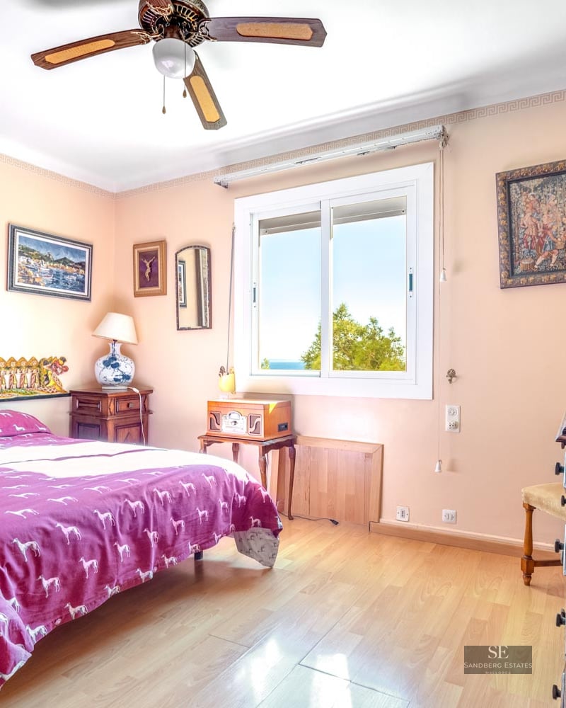 A bedroom featuring a magenta patterned bedspread, peach walls with framed art, wooden floors, and a ceiling fan.