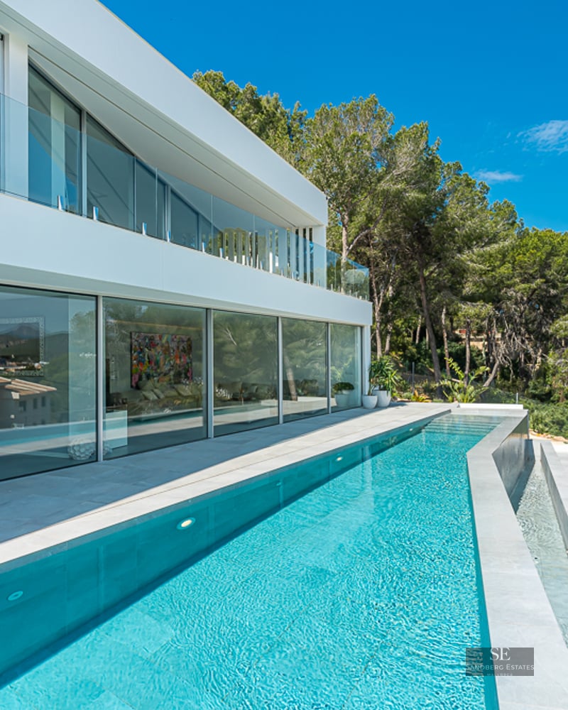 A sleek modern white villa with a long turquoise lap pool, floor-to-ceiling glass windows, and lush green trees.