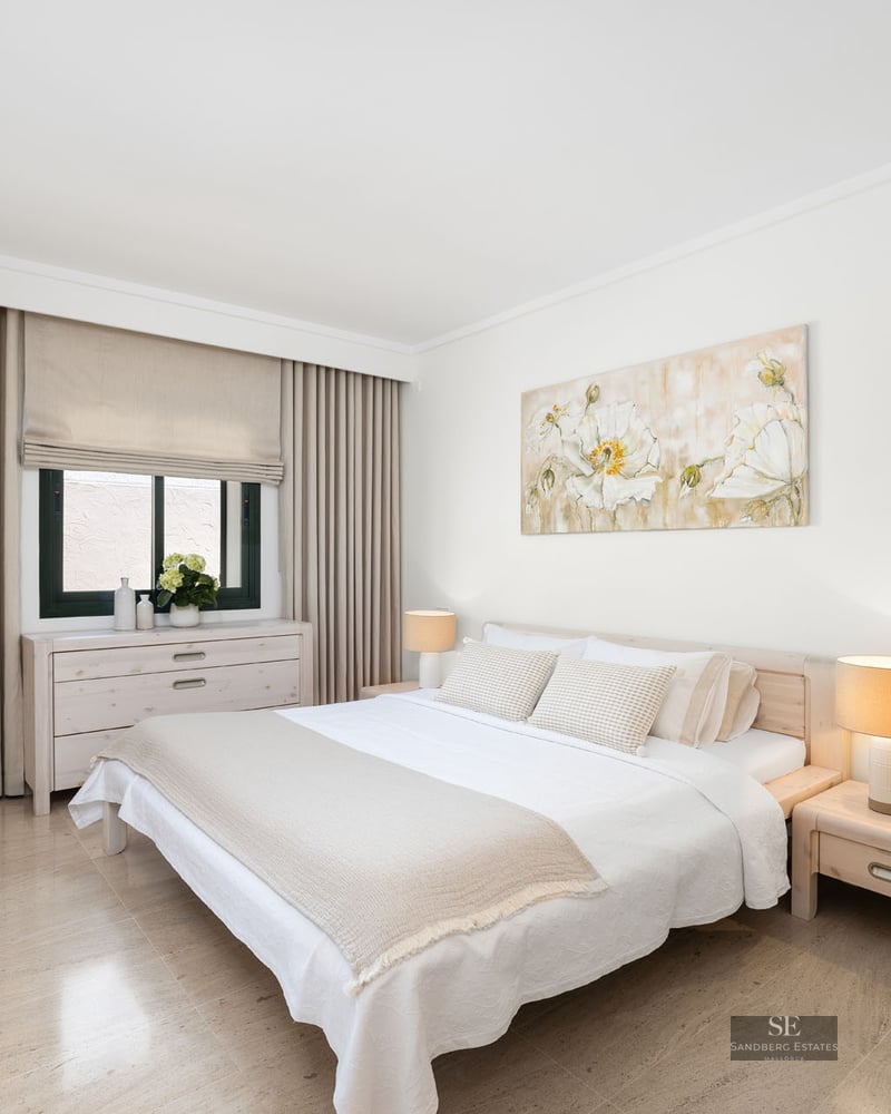 Chambre lumineuse avec lit blanc, tables de chevet en bois, tableau floral et sol en carrelage beige.