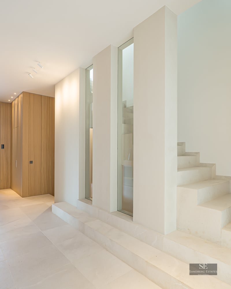 Modern hallway with cream stone floors, a minimalist stone staircase, and light oak wood paneling under bright light.