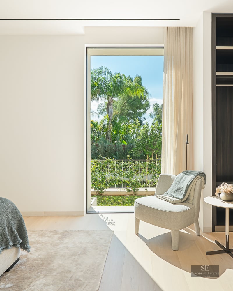 Bright modern bedroom featuring a large dark wood closet, a lounge chair, and a floor-to-ceiling view of a tropical garden.