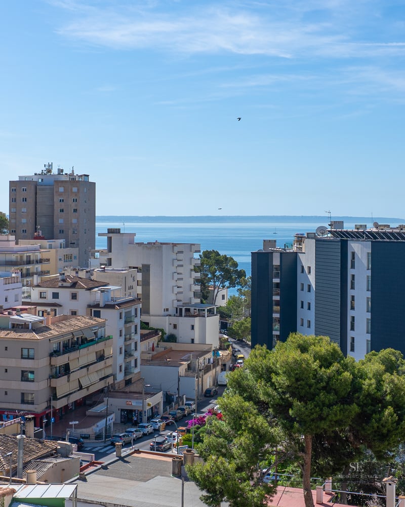 Elevated view of a Mediterranean coastal town with various buildings, green trees, and the blue sea on the horizon.