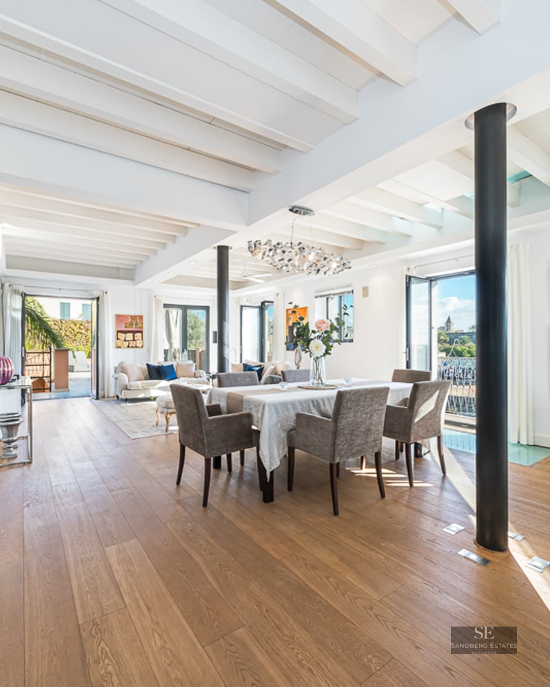 Bright open-plan room with wooden floors, white beamed ceiling, dining set, and a black staircase on the left.