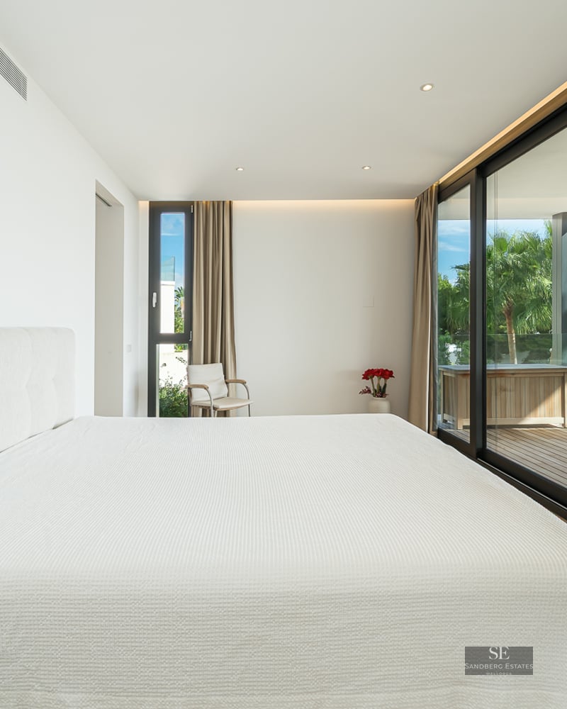 Minimalist white master bedroom with floor-to-ceiling glass doors opening onto a sunny wooden terrace with palm trees.