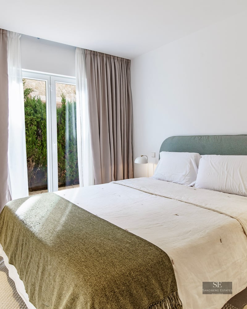 Bright bedroom with large bed, gray headboard, wooden nightstands, and floor-to-ceiling windows with beige curtains.