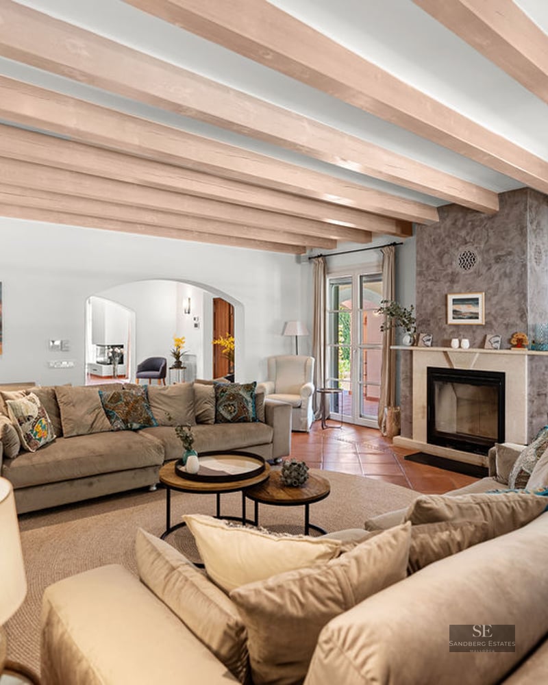 Spacious living room with beige sofas, stone fireplace, wooden ceiling beams, and terrace access.