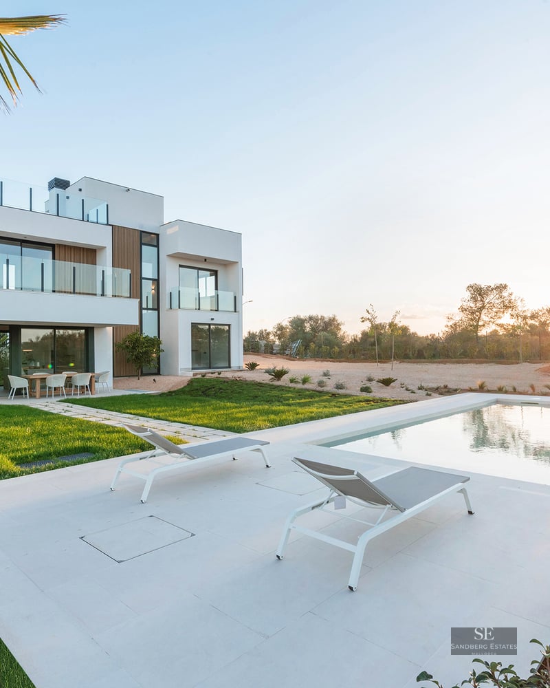 Modern white two-story villa with a swimming pool, sun loungers, and green lawn under a golden sunset sky.