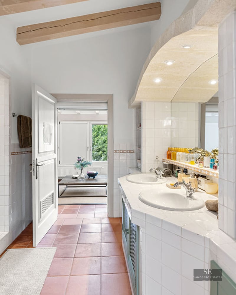 Spacious bathroom with terracotta floors, double white sinks, wooden ceiling beams, and a walk-in tiled shower.