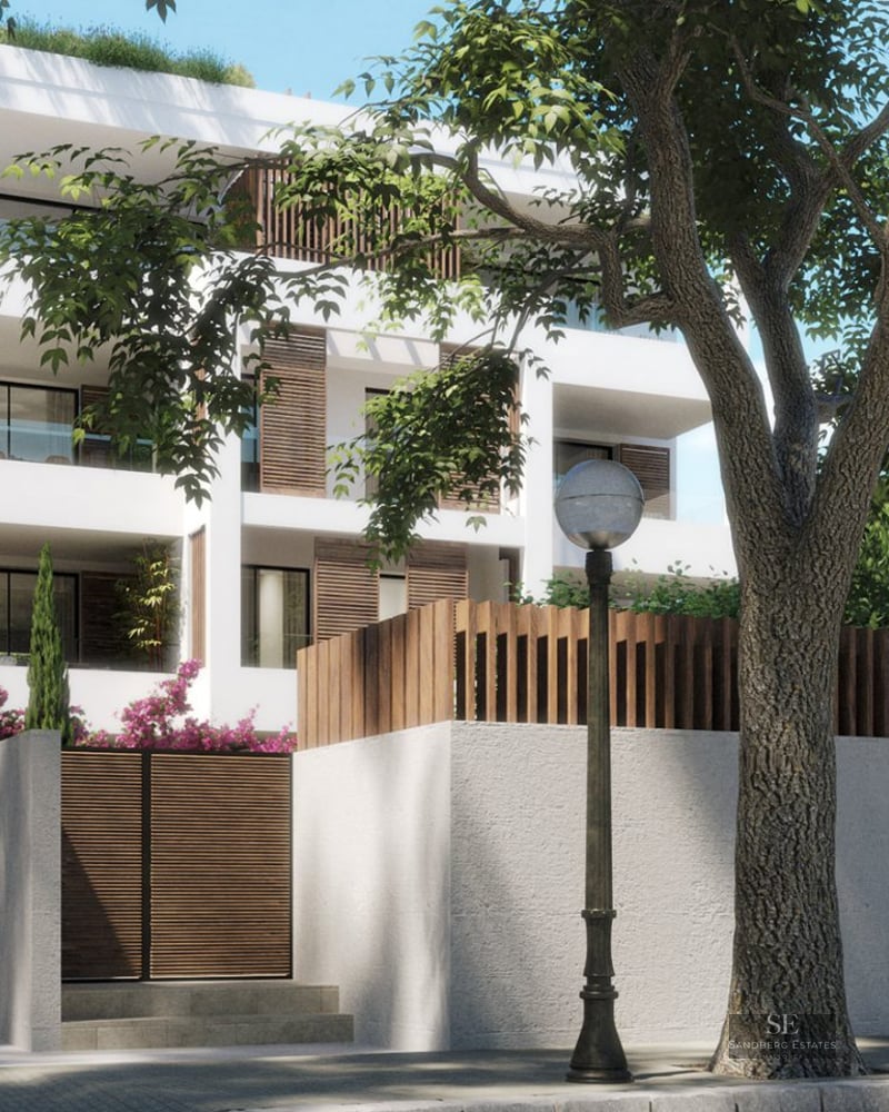 Modern white building with wooden shutters and balconies, behind a wall with a large tree and a blue car in front.