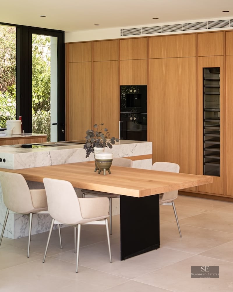 Modern kitchen with wood cabinets, marble island, integrated dining table, and large glass windows showing garden views.