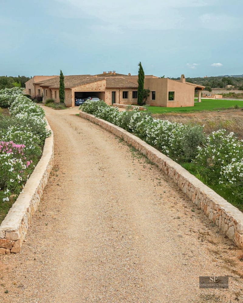 A gravel driveway lined with white oleander flowers leading to a rustic stone villa under a soft blue sky.