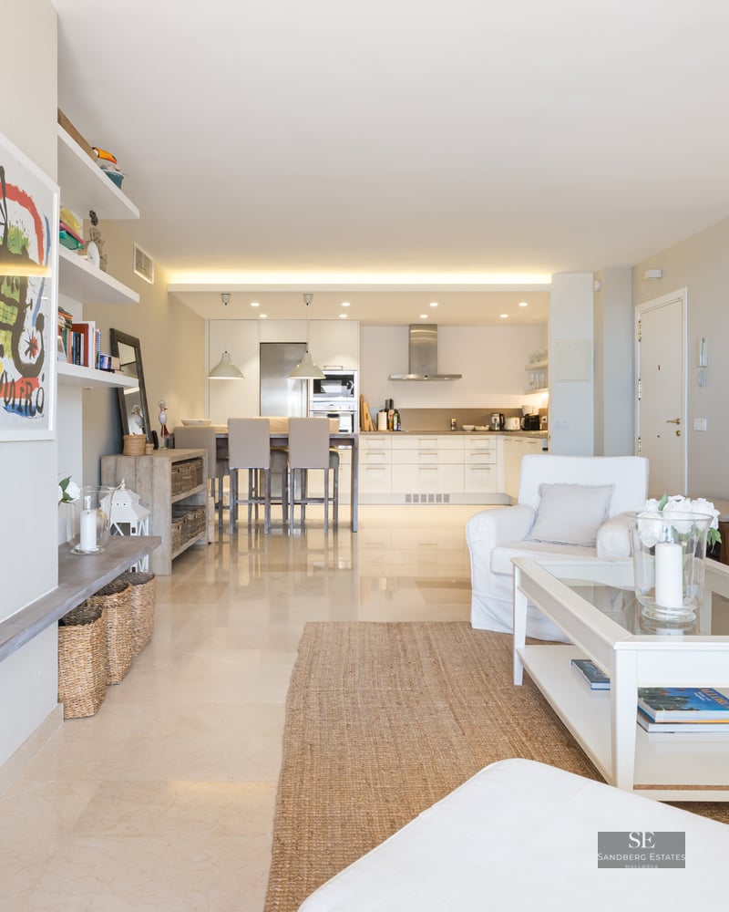 Bright living room with cream sofa, white coffee table, and open kitchen in the background with a breakfast bar.