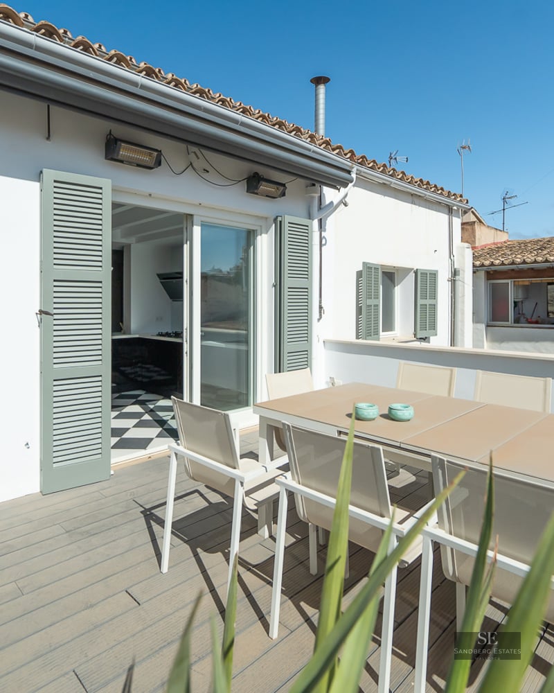 Sunny terrace with a dining table for six, sage green shutters, and wooden decking under a clear blue sky.