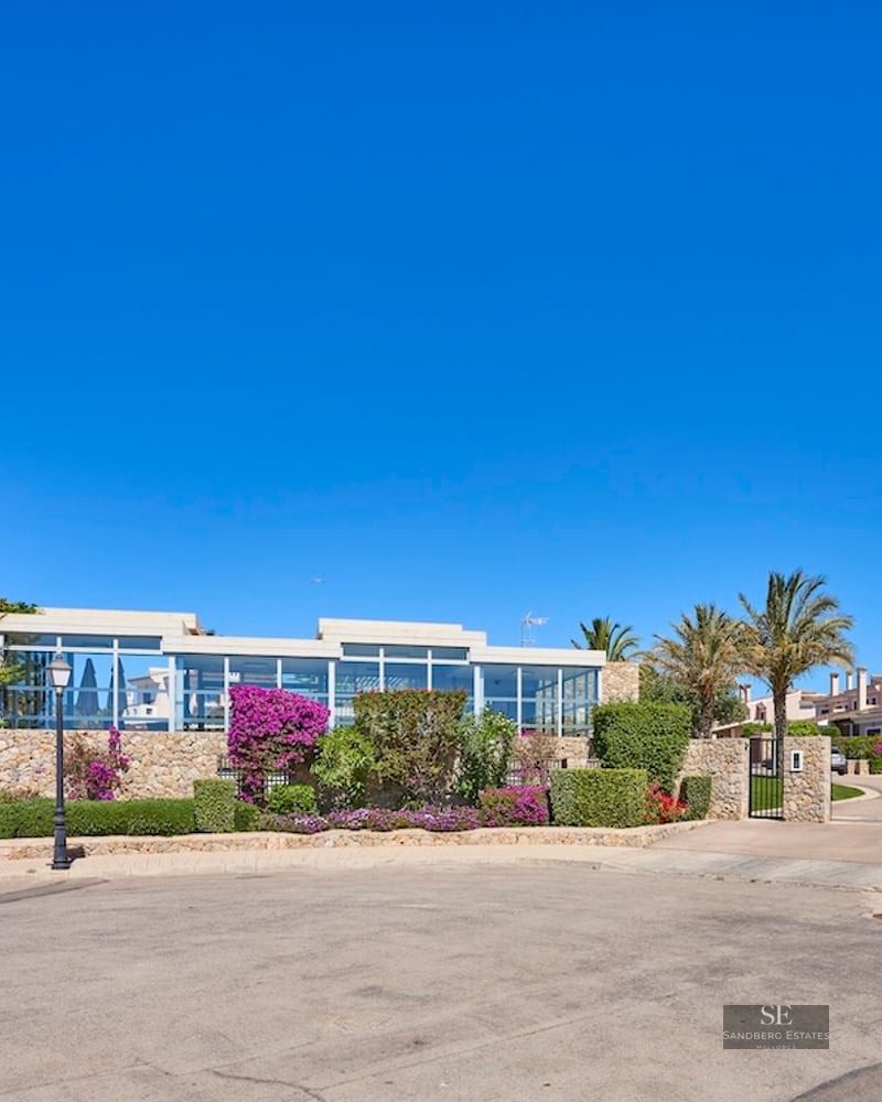 Stone entrance with "Marina Golf" sign under a clear blue sky and Mediterranean landscaping.