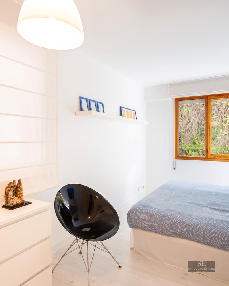 Bright bedroom with a large bed, wooden window looking at greenery, a black designer chair, and a white dresser.