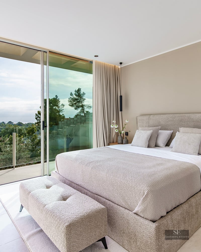 Luxury bedroom with a beige bed and floor-to-ceiling glass doors opening to a balcony with a scenic green view.