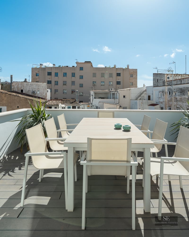 Modern rooftop terrace with white dining set on grey wooden decking overlooking city buildings under a blue sky.