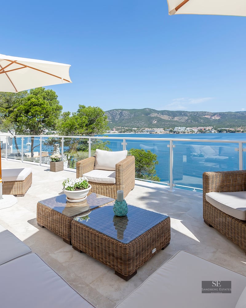 Sunlit terrace with wicker lounge furniture, white umbrellas, and glass railings overlooking a blue sea and mountains.