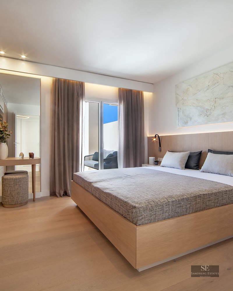 Modern master bedroom with a large wooden bed, integrated lighting, wood paneling, and access to a sunny balcony.