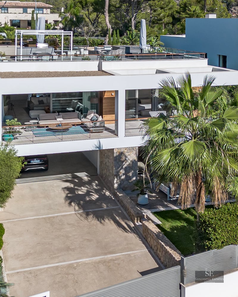 Vue architecturale d'une villa moderne blanche avec terrasses, palmiers et murs en pierre sous une lumière vive.