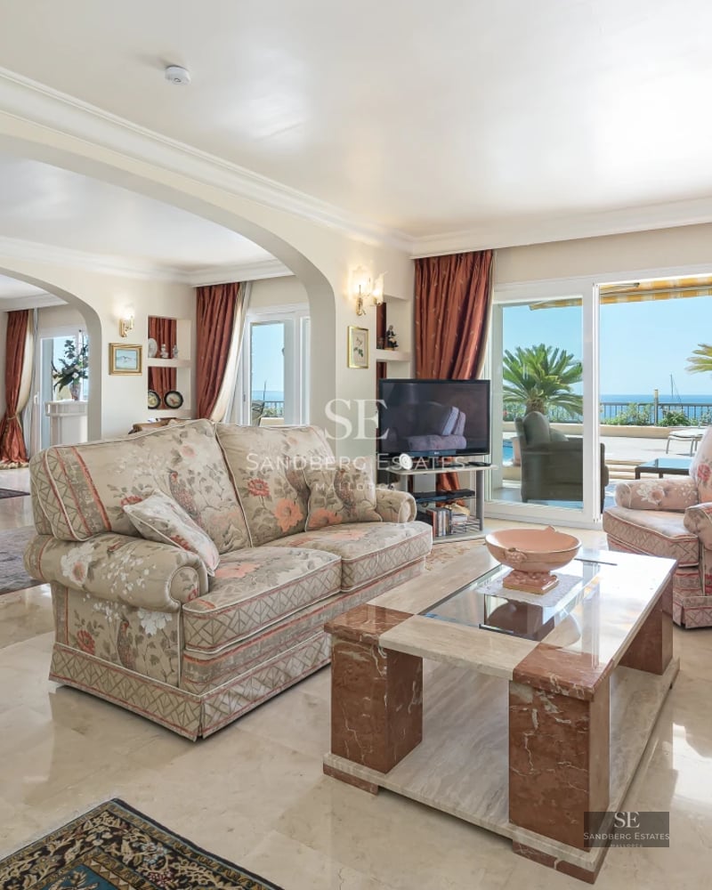 Spacious living room with large windows, light wood floors, and garden view. Modern gray sofa and minimalist decor.