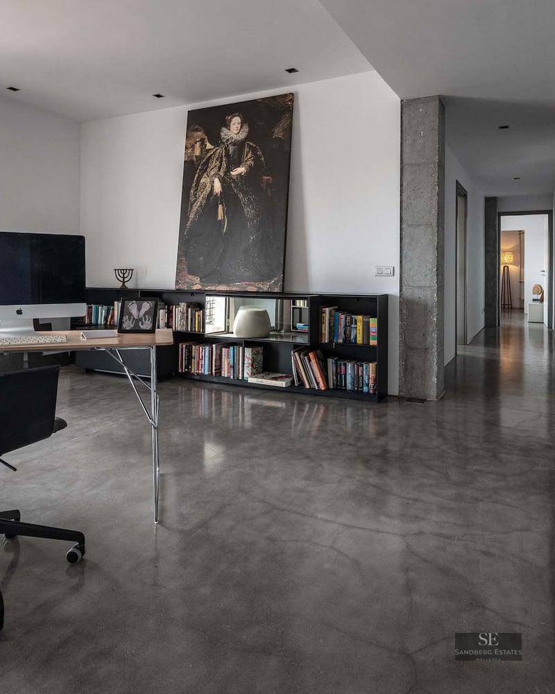 A minimalist home office featuring a desk, iMac, large portrait art, and polished concrete floors leading to a hallway.