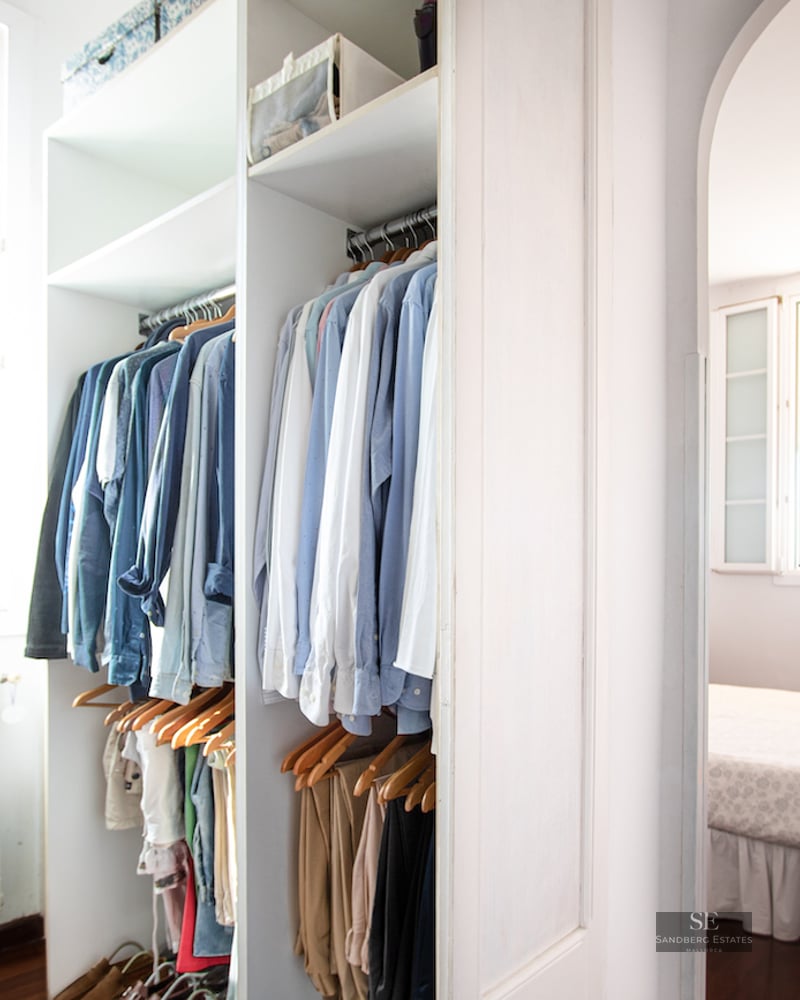 Open white closet with clothes hanging next to a window, looking into a sunlit bedroom.