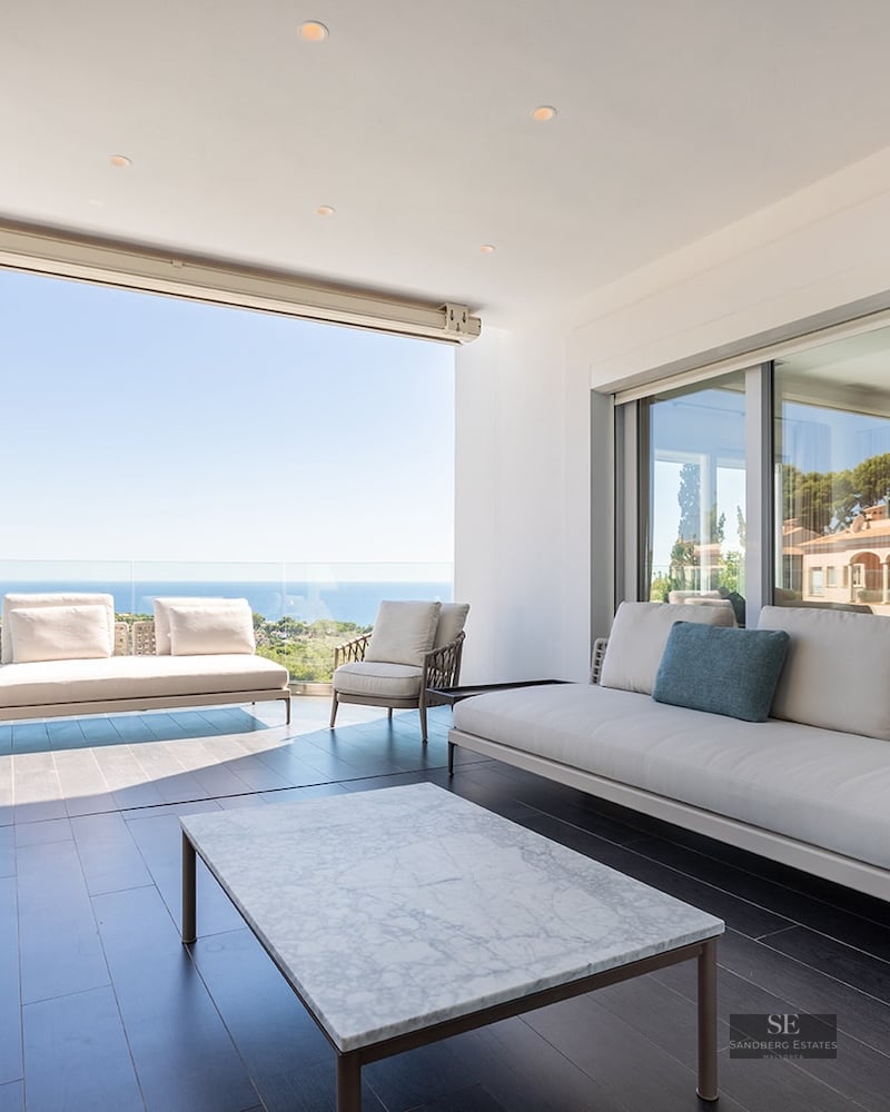 Modern covered terrace with white sofas, a marble coffee table, and a glass railing overlooking the Mediterranean sea.