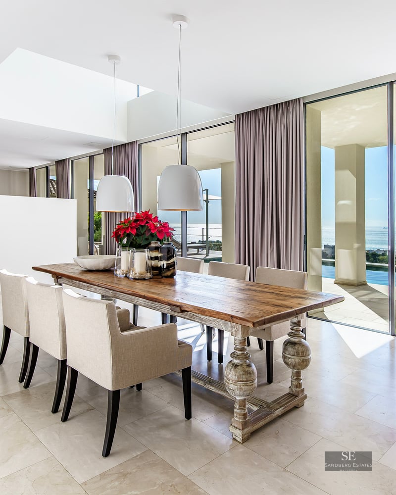 Spacious dining room with a large wooden table, cream chairs, and floor-to-ceiling windows overlooking a sea and pool.
