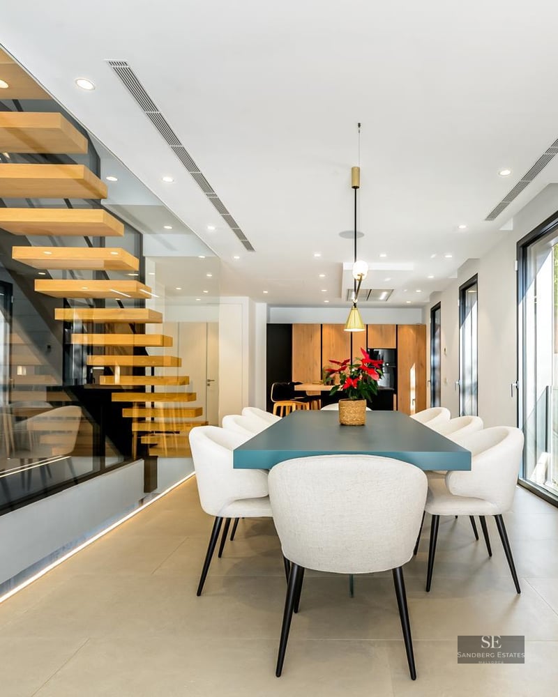 Modern dining room featuring a teal table, white chairs, and a glass-enclosed floating wooden staircase.