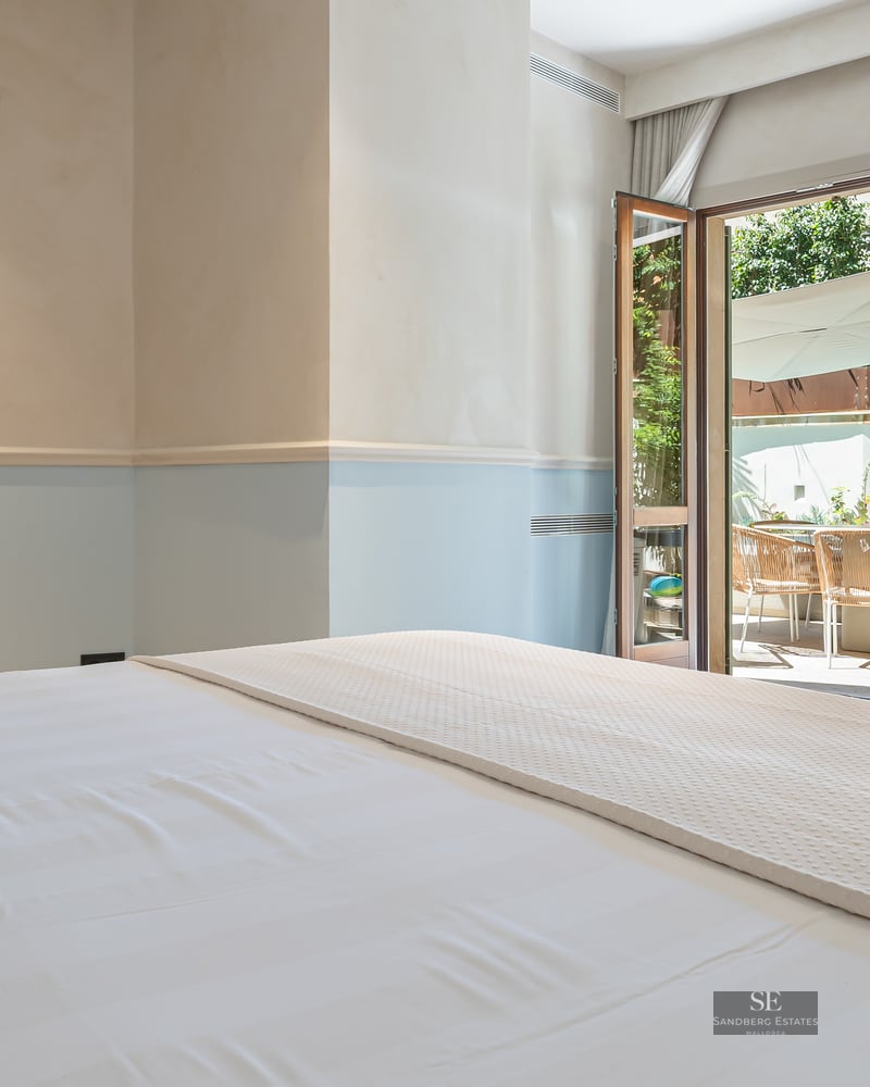Bedroom with white linens and light blue walls looking out onto a sunny terrace through open French doors.