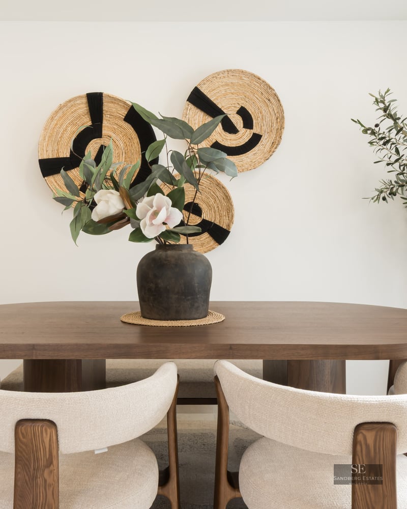 Dining room featuring an oval wooden table, cream chairs, woven wall art, and a large indoor olive tree.