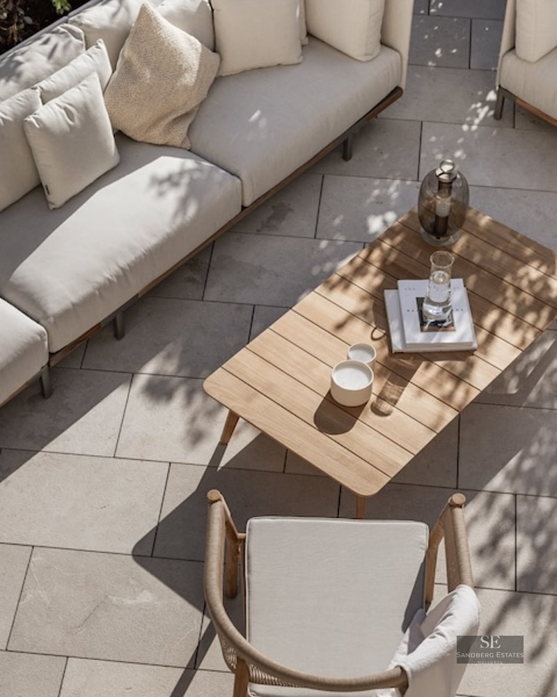High-angle view of a luxury terrace with beige sofas, a wooden coffee table, and stone paving in dappled sunlight.