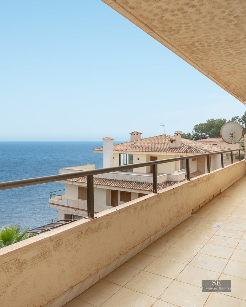A wide tiled balcony with a metal railing overlooking the blue ocean and nearby Mediterranean buildings.