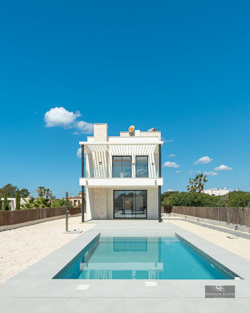A rectangular blue swimming pool leading to a modern white two-story villa under a clear blue sky.