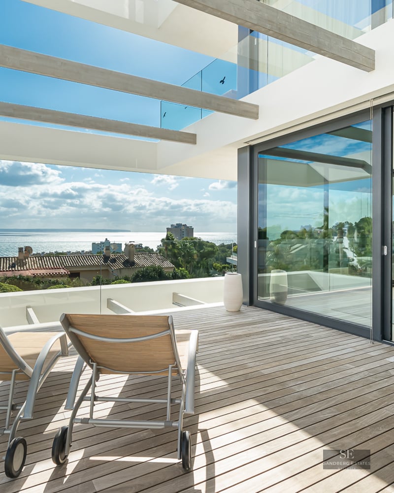 Sun loungers on a wooden terrace with glass railings overlooking the sea, adjacent to an open-plan dining room.