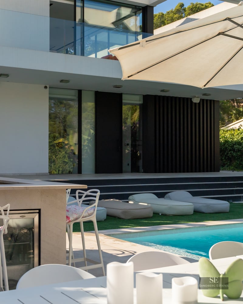 Modern swimming pool with lounge bean bags, outdoor kitchen, and white umbrellas next to a contemporary villa.
